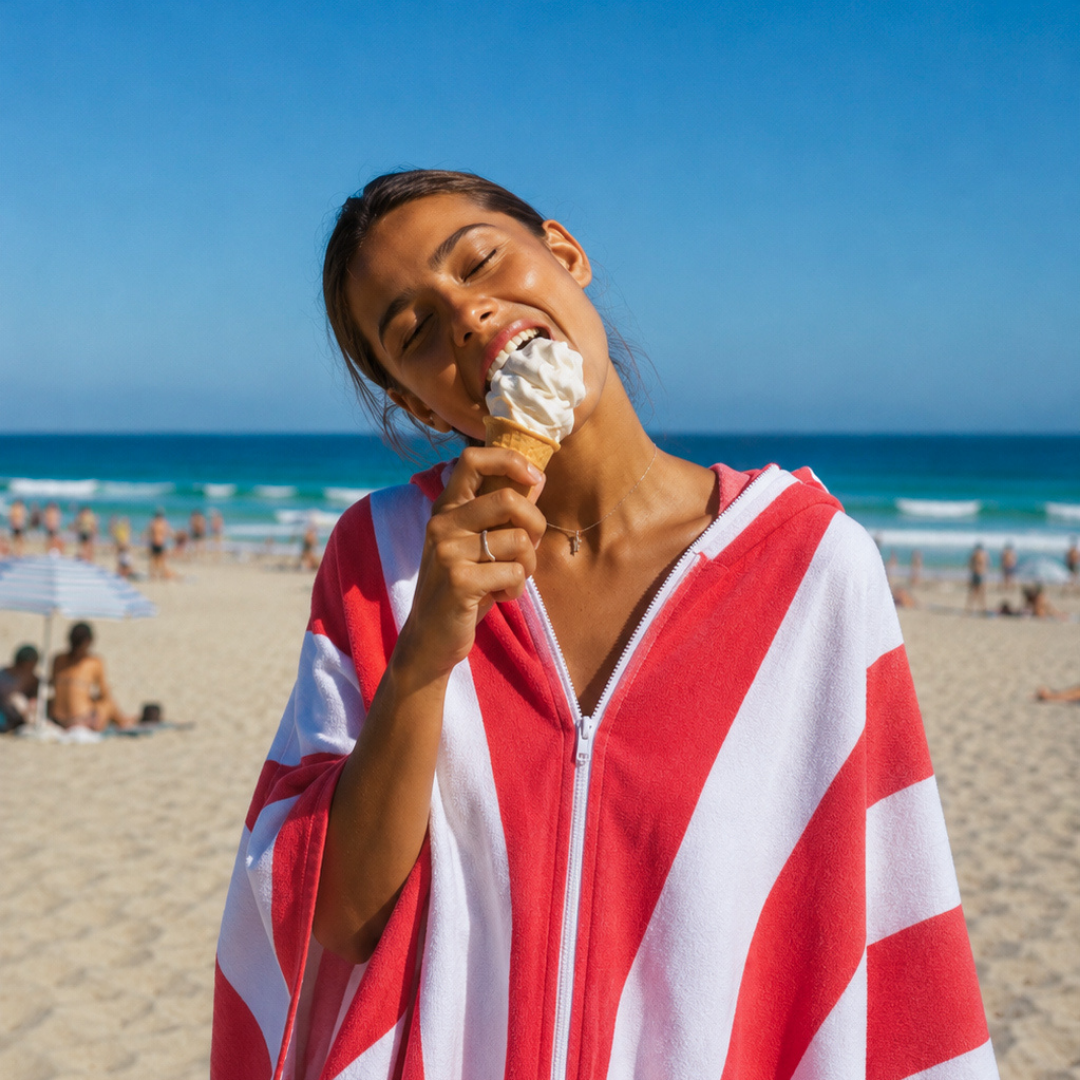 Woman eating an ice cream with both hands while wearing the zipped red and white Zippa microfibre towel on the beach